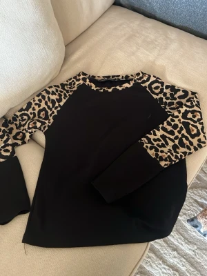 Svart topp med leopardärmar - Cool svart topp med långa ärmar i leopardmönster. Ärmarna och halsringningen har beige och svarta leoparddetaljer som ger en trendig kontrast till den svarta kroppen. Perfekt för dig som vill sticka ut lite extra.