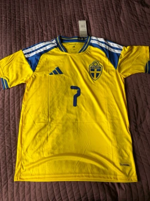 Sverige fotbollströja Bergvall #7 - Gul och blå Sverige fotbollströja från Adidas med nummer 7 och namnet Bergvall på ryggen. Tröjan har korta ärmar, blå detaljer på axlar och ärmslut samt broderat landslagsemblem på bröstet. Tillverkad i lätt och ventilerande material.