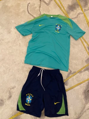 Nike Brasil fotbollsträningsset - Säljer ett Brasilien fotbollsträningsset från Nike. Tröjan är turkos med gröna axelpartier och har Brasil-logga och Nike-swoosh på bröstet. Shortsen är mörkblå med gröna detaljer, snörning i midjan och Brasil-logga samt Nike-märke. Perfekt för träning eller match!