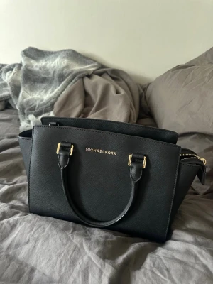 Svart plånbok från Michael Kors och handväska i svart - Snygg svart plånbok från Michael Kors i stilren design med guldfärgade detaljer och dragkedja. Tillverkad i tåligt saffianoläder och har loggan i guld framtill. Perfekt storlek för att få plats med kort, cash och mynt. Enkel att matcha med olika väskor.