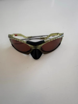 Oakley glasögon  - Säljer ett par sportiga solglasögon från Oakley med grönmelerad båge och bruna linser. Modellen har en futuristisk design med bred näsbrygga och svarta detaljer. Perfekta för utomhusaktiviteter och kommer med originalfodral och mjuk påse.