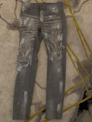 Grå slitna skinny jeans med hål - Säljer ett par grå skinny jeans med slitningar, hål och vita färgstänk. Jeansen har en tight passform och trendiga detaljer som ger en edgy look. Perfekta för dig som gillar streetstyle och vill sticka ut.