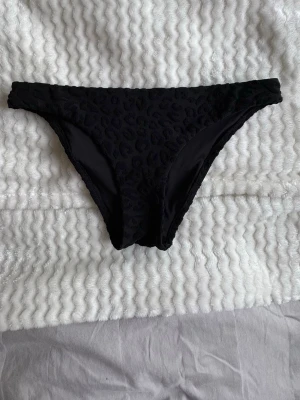 Svarta bikiniunderdel med leopardmönster - Snygg svart bikiniunderdel med diskret leopardmönster i strukturen. Klassisk modell med låg midja och mjukt material som känns bekvämt mot huden. Perfekt för stranddagar eller poolhäng.