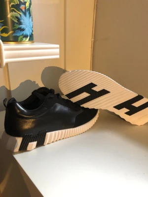 Svarta sneakers med vit sula - Stilrena svarta sneakers med chunky vit sula och svarta detaljer på yttersulan. Ovandelen är i blankt material med en modern, sportig look. Skorna har lågt skaft och snörning framtill, perfekt för en trendig streetstyle.