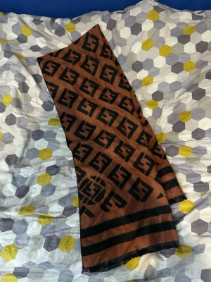 Brun Fendi halsduk med logomönster - Snygg brun halsduk från Fendi med svart logomönster över hela tyget och två svarta ränder i ena änden. Halsduken har rektangulär form och fransar i kanterna. Perfekt för att lyfta din outfit med en lyxig touch.