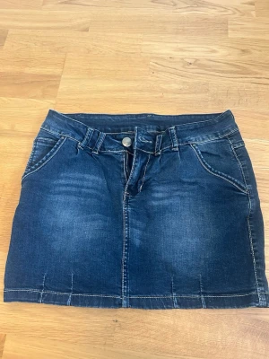 Mörkblå jeanskjol från Flame - Snygg mörkblå jeanskjol från Flame med klassisk femficksdesign, bälteshällor och knapp- samt dragkedjestängning framtill. Kjolen har en kort passform och är tillverkad i denim med tydliga sömmar för en cool look.