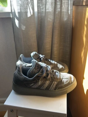 Grå Adidas Samba  - Säljer ett par grå Adidas Samba sneakers i mocka med klassiska tre ränder på sidan och Samba-logga i silver. Skorna har låg profil, rund tå och snörning framtill. Yttersulan är i gummi och ger en retrovibe. Perfekta för dig som gillar streetwear och tidlös stil.