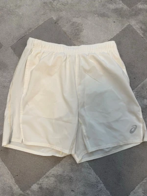 Vita träningsshorts från Asics - Snygga vita träningsshorts från Asics med elastisk midja och dragsko. Shortsen har meshpaneler på sidorna för extra ventilation och en diskret Asics-logga på benet. Perfekta för löpning, gym eller annan sport.