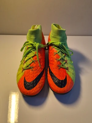 Nike Hypervenom Phantom III FG - Säljer ett par Nike Hypervenom Phantom III FG fotbollsskor i neonorange och limegrönt med svart Nike-logga. Skorna har strumpliknande skaft, snörning och mönstrad ovandel i syntetmaterial för bra grepp och bollkontroll. Perfekta för spel på naturgräs.