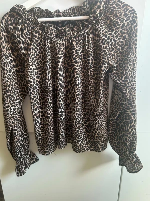 Leopardmönstrad blus från Gina Tricot - Snygg leopardmönstrad blus från Gina Tricot med volangkant vid halsen och långa ärmar med resår och volang vid ärmslut. Blusen har en lös passform och är tillverkad i ett glansigt, mjukt material som ger en lyxig känsla.
