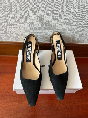 Svarta pumps från Jacquemus - Säljer ett par svarta pumps från Jacquemus med spetsig tå och öppen häl. Skorna har en stilren design i mockaliknande material och bred rem över hälen. Perfekta för dig som gillar minimalistisk och trendig stil.