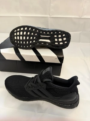 Svarta Adidas Ultraboost sneakers - Snygga helsvarta Adidas Ultraboost sneakers med stickad ovandel och klassiska tre ränder på sidan. Skorna har en bekväm och responsiv Boost-sula samt svart yttersula med grepp. Perfekta för en sportig och stilren look.