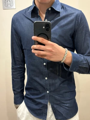 Mörkblå skjorta från Jacob Cohen - Snygg mörkblå denim skjorta från Jacob Cohen med diskreta ränder och klassisk krage. Skjortan har vita knappar och lång ärm, perfekt för en stilren look. Storlek 38 vilket motsvarar en S eller Slim M. Modellen på bilden är runt 190cm och väger 70kg. Nästan helt oanvänd, inga defekter. 