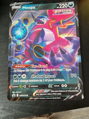 Pokémonkort Hoopa V Fusion Strike - Säljer ett Pokémonkort Hoopa V från Fusion Strike-serien. Kortet har färgstark design med lila, blå och rosa nyanser och visar Hoopa i en dynamisk pose. Kortet har 220 HP och specialförmågan Two-Faced samt attacken Shadow Impact.