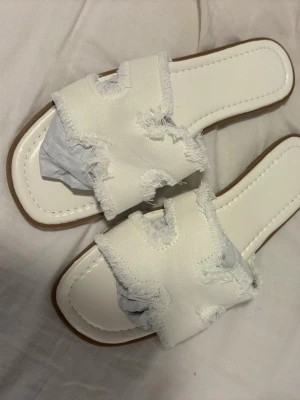 Vita sandaler med fransad kant - Säljer ett par trendiga vita sandaler med breda band i tyg och fransade kanter. Sandalerna har fyrkantig tå och vit sula med kontrastsöm. Perfekta för en avslappnad och stilren look i sommar.