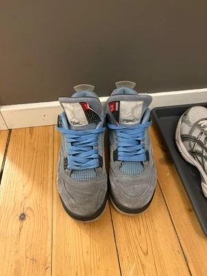 Jordan 4 University Blue sneakers - Snygga Jordan 4 University Blue sneakers med ovandel i ljusblå mocka, meshdetaljer och svarta samt grå inslag på sidorna. Skorna har högt skaft, blå skosnören och klassisk Air-dämpad sula. Perfekta för dig som gillar streetwear och vill sticka ut.
