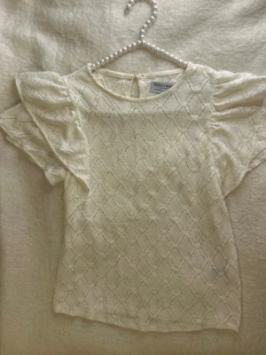 Vit blus med puffärm från - Söt vit blus med korta puffärmar och ett diskret rutmönster i tyget. Blusen har rund halsringning och knapp i nacken. Materialet är lätt och luftigt, perfekt för en fräsch look. Passar till både jeans och kjol. Jätte fin och bra nu till sommaren eftersom den är luftig och sval