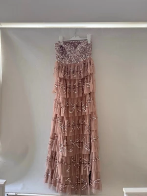 Lång rosa klänning med volanger - Romantisk långklänning i ljusrosa med volanger och broderade blommor. Klänningen är axelbandslös och har flera lager av tunn tyll med vackra detaljer. Använd 1 gång, storlek 38. Säljs för 500 kr.