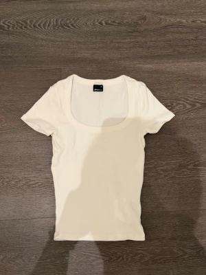 Vit kortärmad topp från Gina Tricot - En stilren vit topp från Gina Tricot med korta ärmar och rund, djup urringning. Tillverkad i mjuk bomull som sitter skönt mot kroppen. Perfekt att matcha med jeans eller kjol för en clean look.