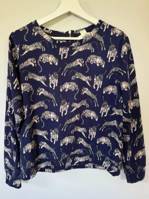 Blå tröja mönstrad med tigrar från H&M, stl S - Blå tröja mönstrad med tigrar från H&M, stl S. Är något nopprig (har inte provat med noppmaskin), annars i fint begagnat skick! Har inga hål eller fläckar vad jag kan hitta. Har haft hund så spår av päls kan förekomma. Rökfritt hem. 
