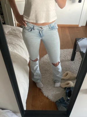 Ljusblå slitna bootcut jeans - Säljer ett par ljusblå bootcut jeans med slitningar vid knäna och snygga broderade detaljer på bakfickorna. Jeansen har låg midja och klassisk femficksdesign. Perfekta för en avslappnad och trendig look.