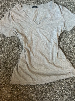 Grå v-ringad t-shirt Tommy Hilfiger - Snygg grå t-shirt från Tommy Hilfiger med v-ringning och korta ärmar. Tillverkad i mjuk bomull och har en liten broderad logga nere vid fållen. Perfekt basplagg till garderoben.