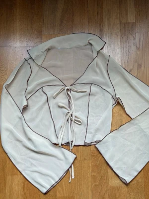 Smörgult mesh topp med snörning - Trendig smörgul mesh topp med långa ärmar och markerade sömmar i brun kontrast. Toppen har snygg snörning framtill och är croppad med en lätt transparent känsla. Perfekt för dig som gillar en edgy och unik stil.