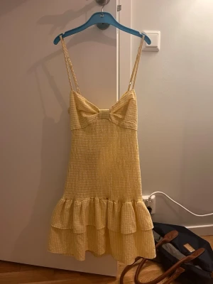 Gul rutig volangklänning med rosett - Söt gul och vit rutig kortklänning med volangkant och stor rosett framtill. Klänningen har smala axelband och omlott-detalj vid bysten. Perfekt för sommarens varma dagar. Från Zara