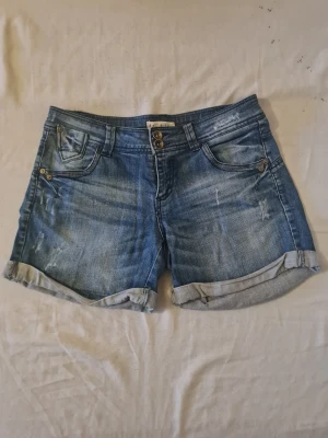 Blå jeansshorts med uppvik - Snygga blå jeansshorts med uppvikta benslut och dubbla knappar i midjan. Klassisk femficksmodell med slitna detaljer för en avslappnad look. Perfekta för varma dagar och enkel att matcha med olika stilar.