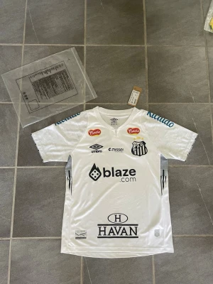Vit Santos FC fotbollströja från Umbro - Snygg vit fotbollströja från Santos FC med flera tryckta sponsorer som blaze.com och Havan. Tröjan har korta ärmar, rund hals och klubbmärke på bröstet. Tillverkad i lätt och ventilerande material, perfekt för fotboll eller träning.