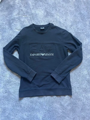 Emporio Armani tröja - Snygg svart sweatshirt från Emporio Armani EA7 med rund halsringning och stort logotryck på bröstet. Tröjan har långa ärmar och ribbade muddar vid ärmslut och nederkant. Perfekt för en stilren och avslappnad look. Priset är inte hugget i sten!