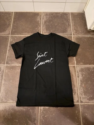 Svart Saint Laurent t-shirt - Snygg svart t-shirt från Saint Laurent med vit logga i handskriven stil på bröstet. Klassisk passform och rund halsringning. Perfekt till jeans eller shorts för en clean och trendig look. Är ca 182 och väger 66