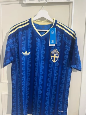 Blå Sverige fotbollströja Adidas - Säljer en blå fotbollströja från Adidas med Sveriges landslagsemblem på bröstet och gula detaljer. Tröjan har ett retroinspirerat mönster, korta ärmar och V-ringning. Tillverkad i lätt och ventilerande material, perfekt för match eller träning.