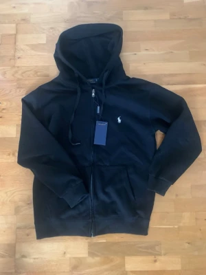 Svart hoodie från Polo Ralph Lauren - Svart hoodie med dragkedja från Polo Ralph Lauren. Klassisk design med huva och dragsnören, två fickor framtill och den ikoniska broderade loggan på bröstet. Perfekt för en chill och stilren look.
