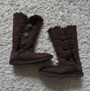 Bruna UGG-stövlar med knappar - Mysiga bruna UGG-stövlar i mocka med tre stora dekorativa knappar på sidan och fluffigt foder. Stövlarna har en grov sula och är perfekta för kalla dagar. Klassisk UGG-design med synliga sömmar och mjukt material.