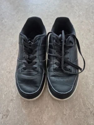 Sneakers - Svarta sneakers från nonation med klassisk låg profil och vita sulor. Skorna har svarta skosnören, diskreta sömmar och ovandel i syntetmaterial. Perfekta för en stilren och enkel look.