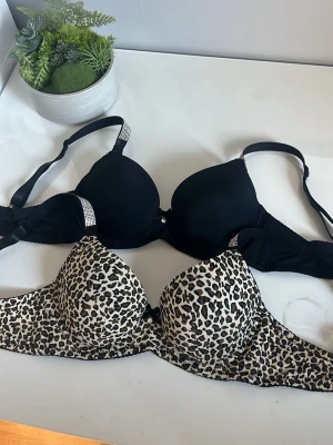 2-pack bhar svart & leopardmönstrad - Säljer två bhar, en i helsvart med glittriga detaljer på axelbanden och en med leopardmönster i beige, svart och brunt. Båda har vadderade kupor och justerbara axelband. Perfekta för dig som vill ha både klassiskt och trendigt i garderoben.