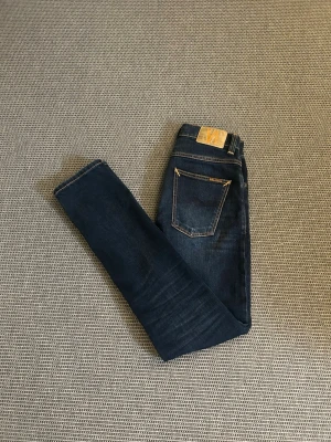 Nudie jeans - Säljer ett par mörkblå jeans från Nudie Jeans med klassisk femficksdesign och kontrastsömmar. Jeansen har en smal passform och är tillverkade i slitstark denim med Nudie-logga på bakfickan och läderpatch i midjan.