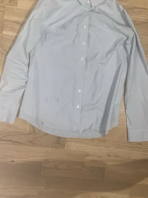 Sail Racing Skjorta - Klassisk ljusblå skjorta med tunna vita ränder och knappar framtill. Skjortan har lång ärm och en liten broderad logga på bröstet. Perfekt till jeans eller chinos för en clean look.