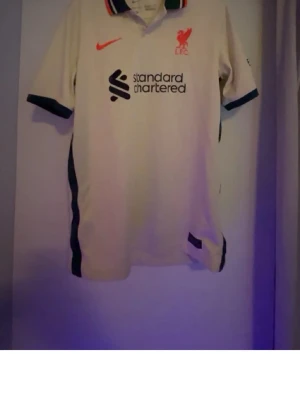 Liverpool FC vit matchtröja - Snygg vit Liverpool FC matchtröja från Nike med röd logga och klubbmärke på bröstet. Tröjan har mörkblå detaljer på krage och ärmslut samt sponsortryck på framsidan. Perfekt för dig som älskar fotboll och vill visa ditt stöd för laget. Köpt på unisport storlek XS