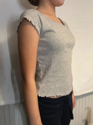 Topp med volangkant  - Säljer en ljusgrå ribbad topp med korta ärmar och volangkant vid ärmslut och nederkant. Toppen har en rundad halsringning och sitter tight mot kroppen. Perfekt till jeans eller kjol för en enkel och snygg look. 😍☀️