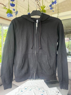 Svart zip hoodie med fickor - En klassisk svart hoodie med dragkedja framtill och två stora fickor. Tröjan har huva med dragsko och ribbade muddar vid ärmslut och nederkant. Perfekt för en avslappnad stil och enkel att matcha med det mesta.