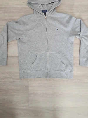 Grå zip hoodie från Polo Ralph Lauren - Klassisk grå hoodie med dragkedja från Polo Ralph Lauren. Hoodien har två fickor framtill, justerbar huva och den ikoniska mörkblå loggan broderad på bröstet. Tillverkad i mjuk bomullsblandning för en skön och avslappnad look. Storlek XL använde den som 13-14 åring. Pris kan diskuteras, hör gärna av er!