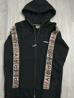 Svart Burberry dress  - Svart hoodie från Burberry med dragkedja framtill och huva med snören. Klassiska Burberry-ränder och logga längs ärmarna samt liten vit Burberry-logga på bröstet. Tillverkad i mjukt material som känns lyxigt och bekvämt. Perfekt för en clean och stilren look.