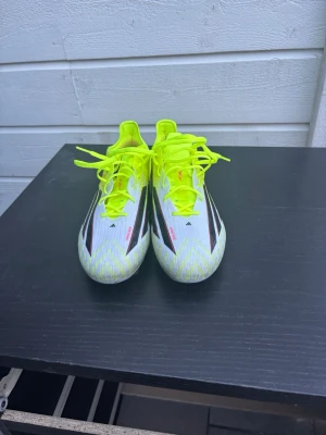 Adidas X Speedflow fotbollsskor - Säljer ett par Adidas X Speedflow fotbollsskor i vitt, svart och neon-gult med röda detaljer. Skorna har snörning, låg profil och mönstrad ovandel i syntetmaterial. Sulan har fasta dobbar för spel på gräs. Perfekta för dig som vill ha lätta och snabba skor på planen.