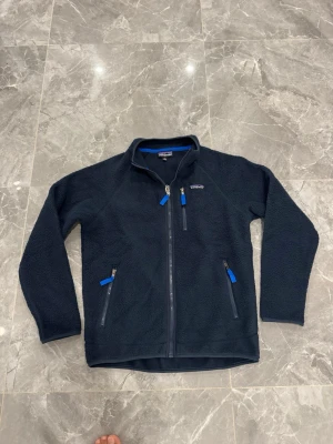Marinblå fleecejacka från Patagonia - Säljer en marinblå fleecejacka från Patagonia med dragkedja framtill och tre fickor med blå detaljer. Jackan har hög krage och är tillverkad i mjukt fleecematerial, perfekt för kyliga dagar. Klassisk och sportig design som funkar till det mesta.