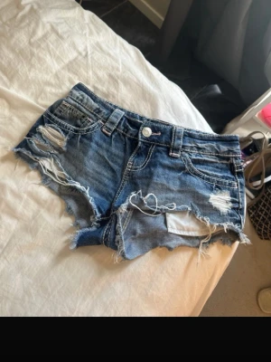 Blå slitna jeansshorts med fransar - Säljer ett par blå jeansshorts med slitna detaljer och fransig kant. Shortsen har fem fickor, bälteshällor och råa kanter för en edgy look. Perfekta för sommaren och ger en avslappnad vibe. Lite stora i storleken 