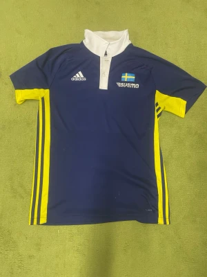Blå-gul Adidas SUEMO sporttröja - Snygg blå sporttröja från Adidas med gula detaljer och vit krage. Tröjan har SUEMO-tryck och svensk flagga på bröstet samt klassiska Adidas-ränder längs sidorna. Perfekt för träning eller sportevenemang.