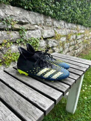Adidas Predator blå/gula fotbollsskor - Säljer ett par Adidas Predator fotbollsskor i svart, blått och gult med högt skaft och snörning. Skorna har de klassiska tre ränderna i vitt och gula detaljer på ovandelen samt gula dobbar. Perfekta för dig som vill ha grym kontroll på planen.