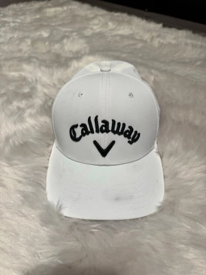 Vit keps från Callaway - Snygg vit keps från Callaway med broderad svart logga framtill och texten 'CHROME TOUR' på sidan. Klassisk böjd skärm och justerbar passform. Perfekt för golfbanan eller som sportig accessoar. Tillverkad i ett lätt och ventilerande material.